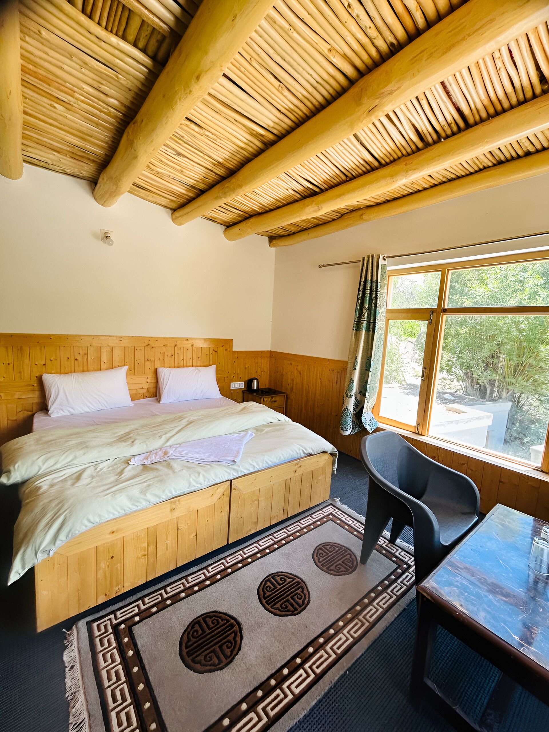 A room with a wooden bed, a table, and a window with a view of greenery.