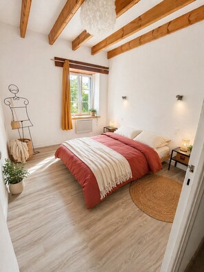 A bedroom with a bed, wooden beams, a window with curtains, a nightstand, and a wall-mounted light fixture. - Charming house * Penty Breton * private spa * enclosed garden * beaches within walking distance (Lampaul-Plouarzel)