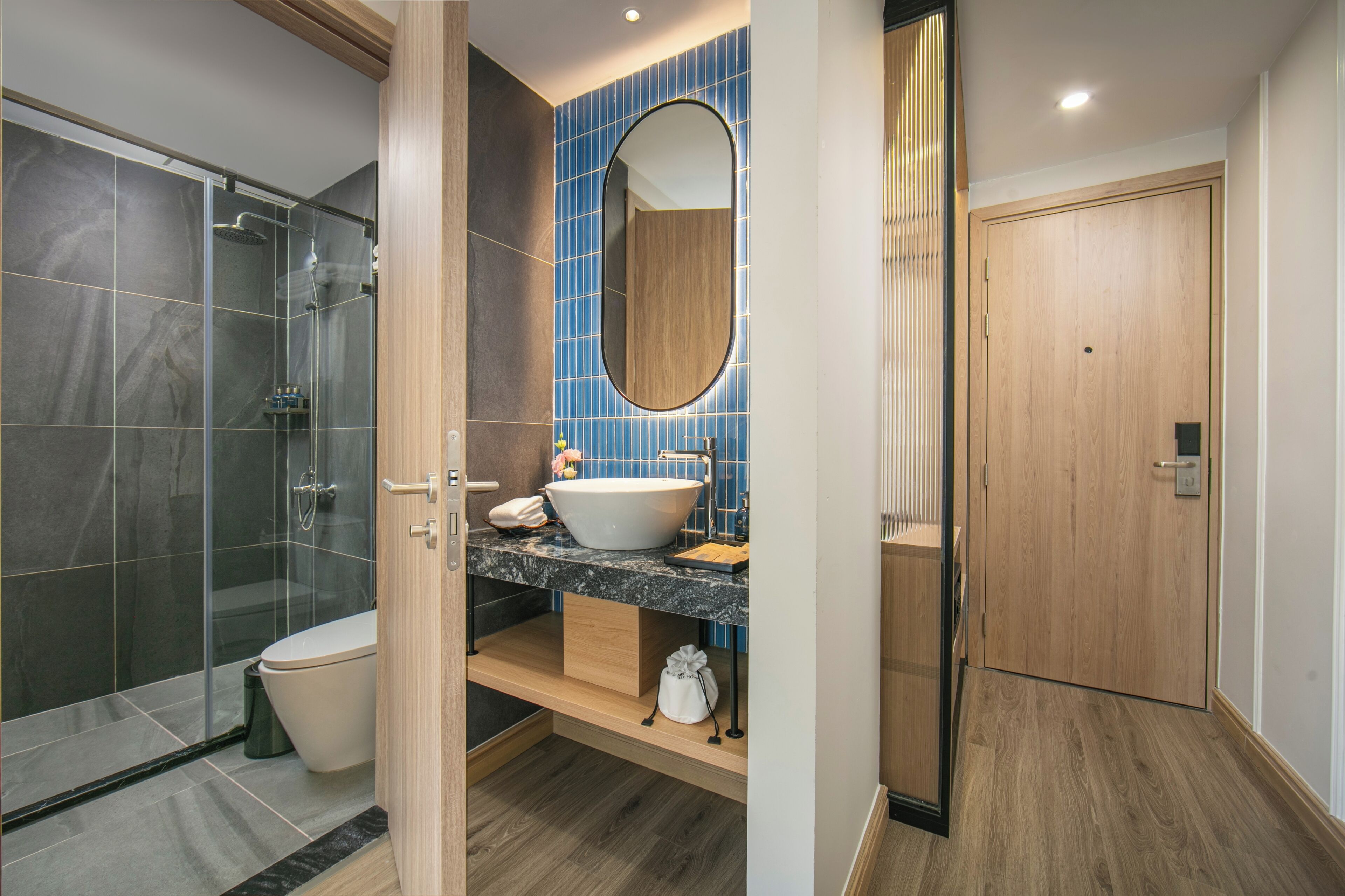 A modern bathroom with a glass shower enclosure, a white toilet, a vanity with a blue-tiled backsplash, and a wooden door.