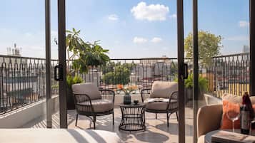 A balcony with two wicker chairs, a small round table, and a sofa. There is a view of a cityscape and a plant in the background.