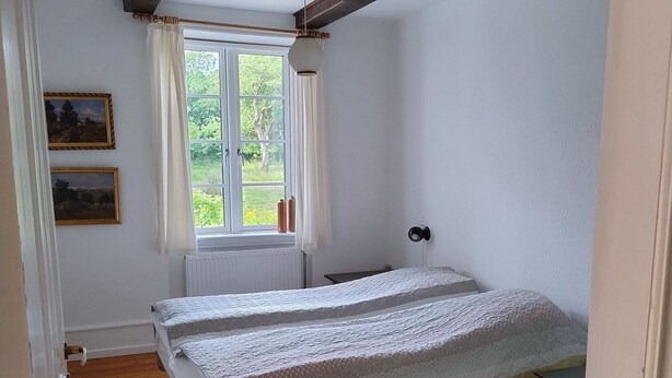 Ein Schlafzimmer mit Holzboden, einem Bett mit weißer und grauer Decke, einem Fenster mit Vorhängen und einem Heizkörper.