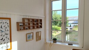Ein Zimmer mit Holztisch, Heizkörper, einem gerahmten Vogelbild, einem Fenster mit Blick auf Grünflächen und einer kleinen Pflanze im Fensterbrett.