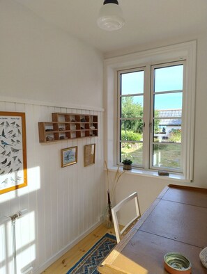 Ein Zimmer mit Holztisch, Heizkörper, einem gerahmten Vogelbild, einem Fenster mit Blick auf Grünflächen und einer kleinen Pflanze im Fensterbrett.