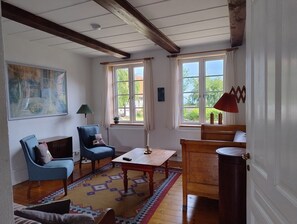 A room with a wooden floor, a blue armchair, a wooden table, and a window with a view of greenery. - Bornholm – Lovely apartment with sea view (Hollændergårde)