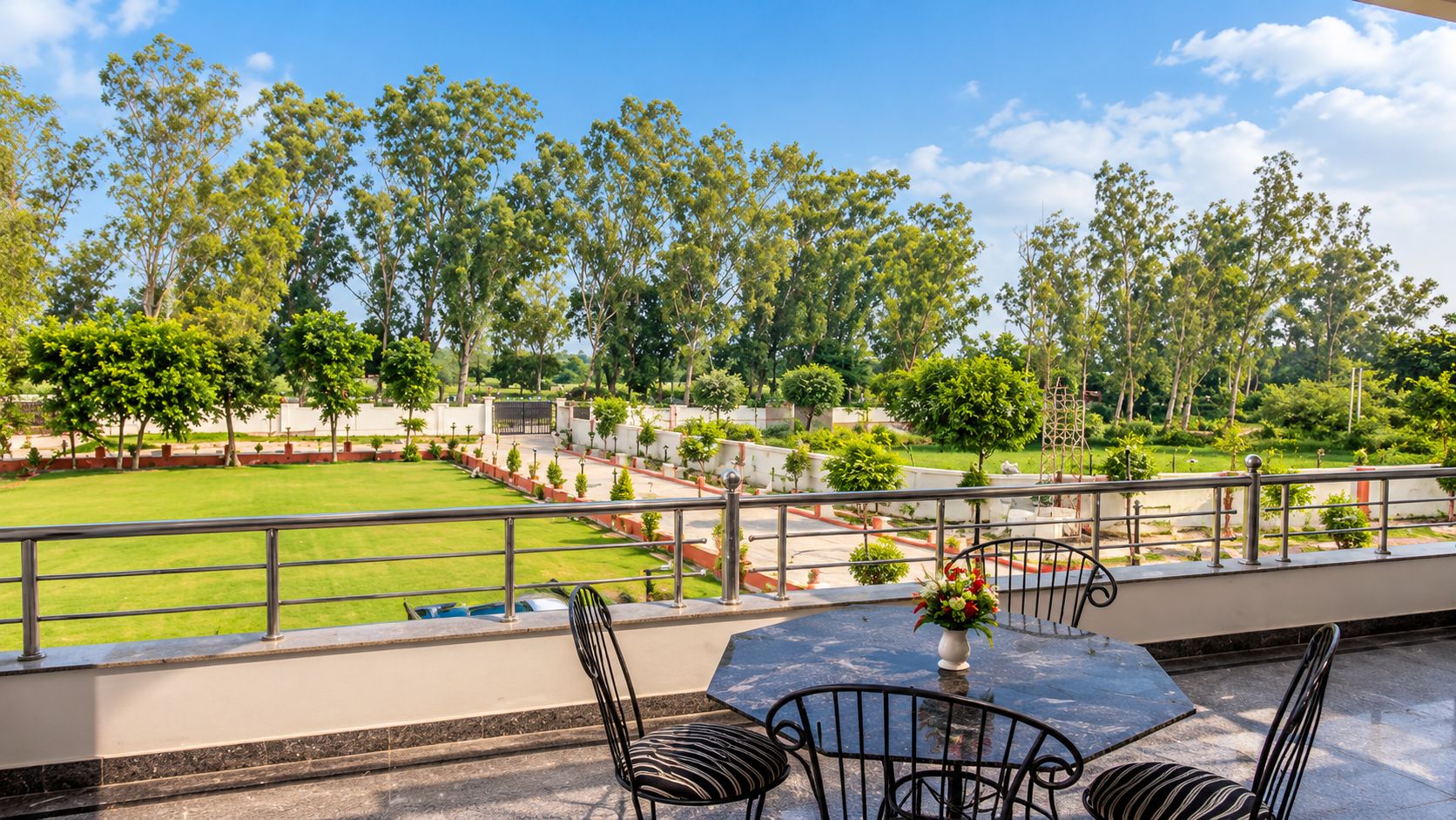 A terrace with a view of a well-maintained lawn and trees, a table with chairs, and a decorative vase.