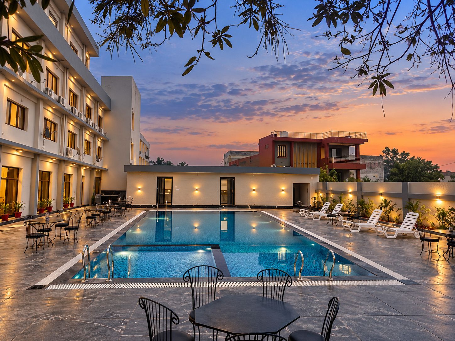 A swimming pool with lounge chairs and tables, surrounded by buildings with balconies and outdoor lighting.