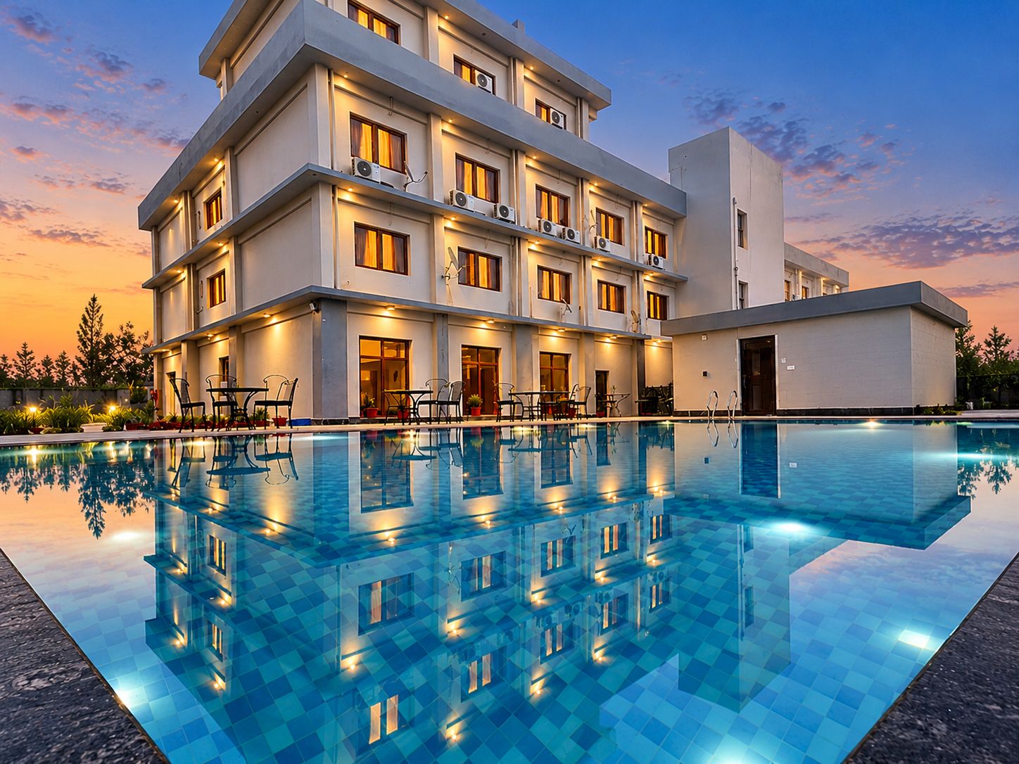 A multi-story hotel with a swimming pool, outdoor seating, and a sunset sky.
