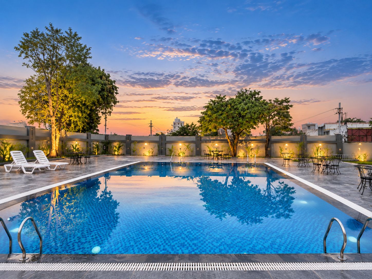 A rectangular swimming pool with a tiled edge, surrounded by lounge chairs and tables, with trees and a sunset in the background.