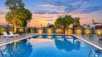 A rectangular swimming pool with a tiled edge, surrounded by lounge chairs and tables, with trees and a sunset in the background.