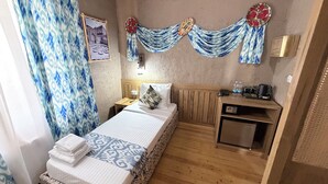 Une petite chambre avec un lit simple, une tête de lit en bois, une table de chevet avec un téléphone et une bouilloire, et une petite armoire.