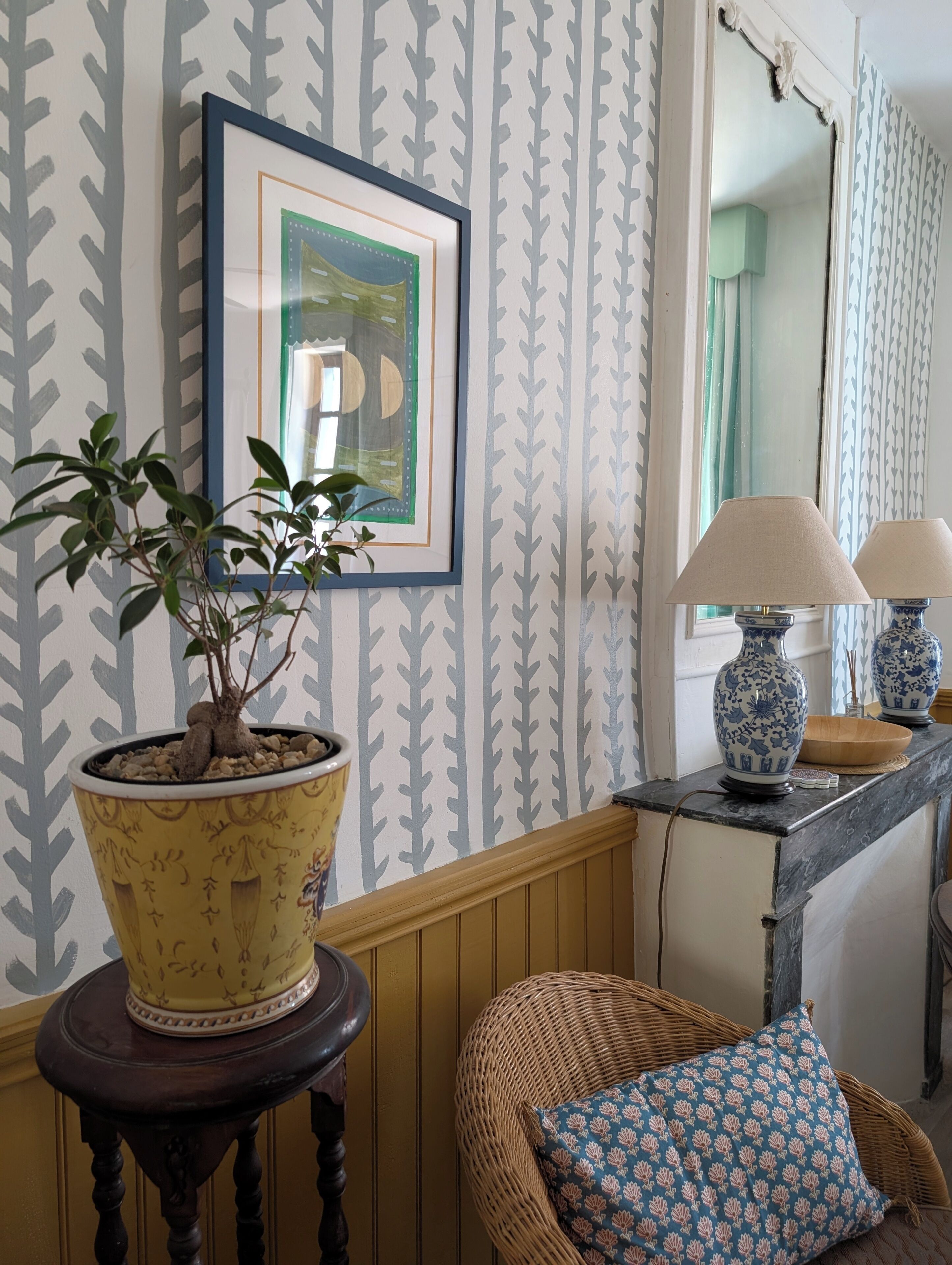Een kamer met behang met een patroon, een ingelijst schilderij, een kamerplant, een rieten stoel met kussen, en twee lampen op een tafel.