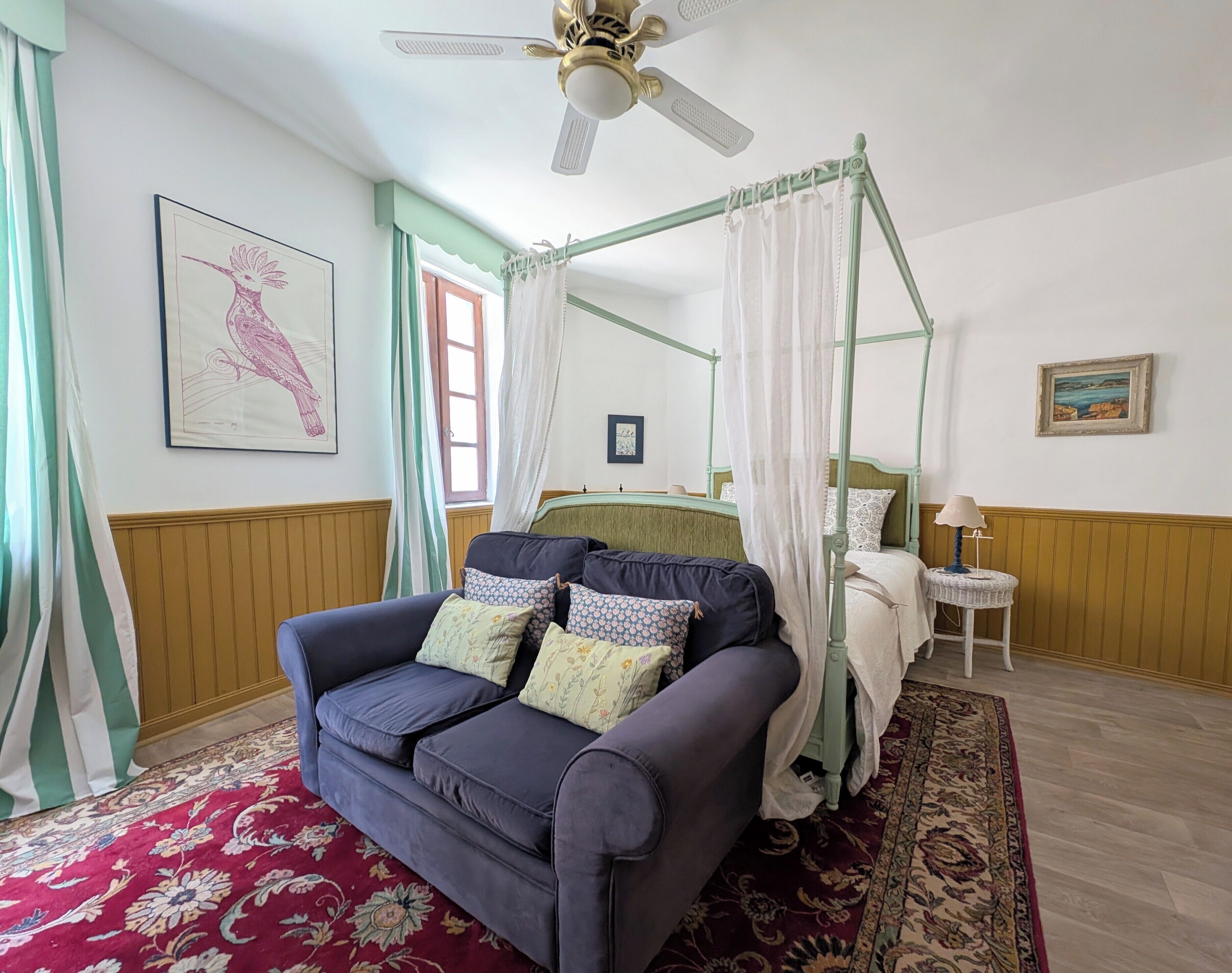 A room with a four-poster bed, a ceiling fan, a floral-patterned sofa, and a red carpet.