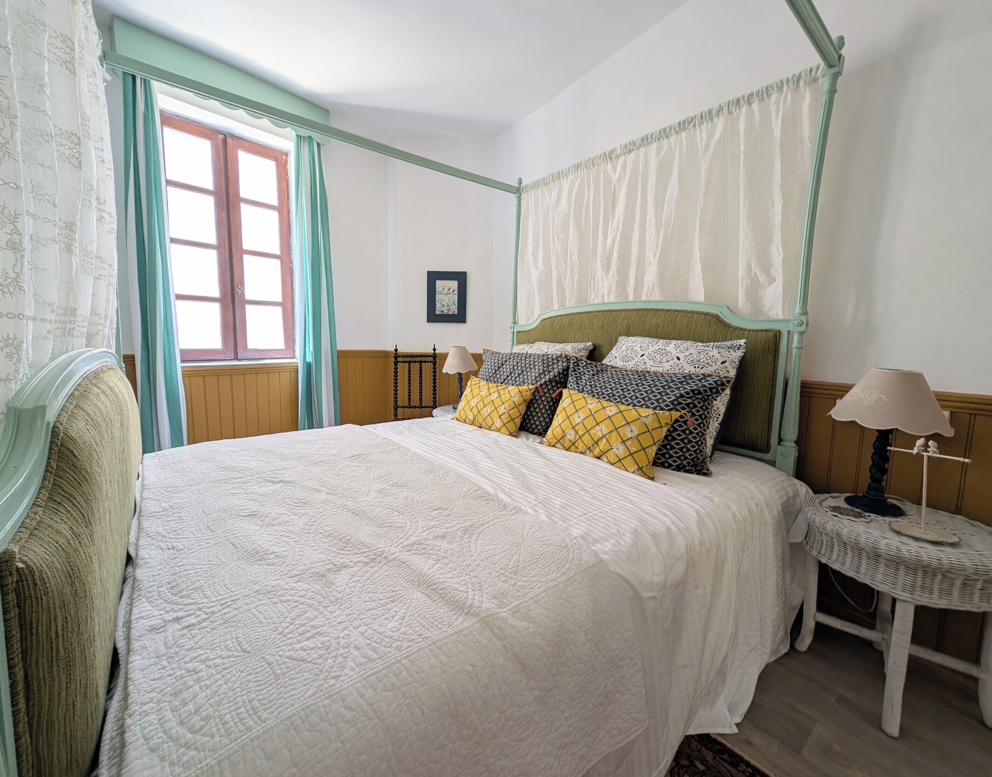 A bedroom with a four-poster bed, a bedside table with a lamp, and a window with curtains.