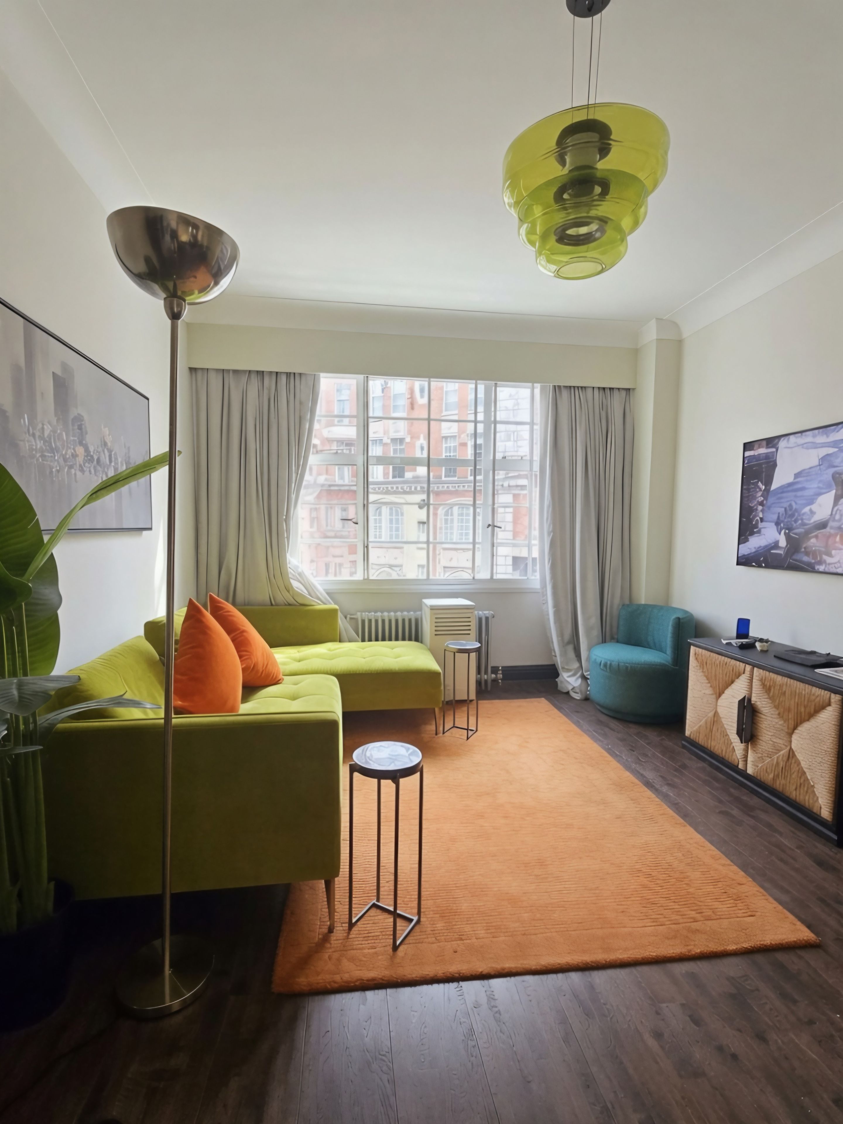 A modern living room with a green sofa, a small side table, a floor lamp, a teal armchair, and a large window with curtains.
