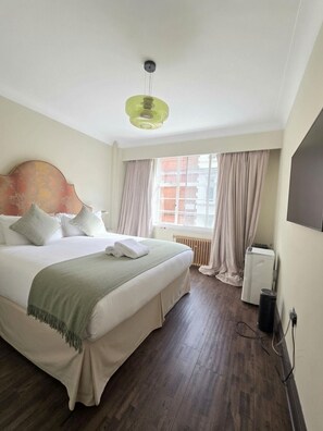 A hotel room with a large bed, a headboard, a window with curtains, a radiator, and a wall-mounted TV. - Prime 2 bedrooms flat in Knightsbridge- Harrods view / portable air conditioning (London)