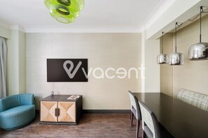 A modern hotel room with a dining area, a blue chair, a wooden cabinet, and a wall-mounted TV displaying the logo 'vacant'. - Prime 2 bedrooms flat in Knightsbridge- Harrods view / portable air conditioning (London)