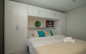 A bedroom with a bed, white wardrobe, and a shelf with decorative items. - Corporate Stays First Living - 206 (Asunción)