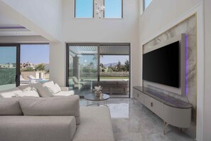 A modern living room with a large TV, a sofa, a coffee table, and a glass door leading to an outdoor area. - Atriora Luxury Villa | Private Pool & 150m to sea! (Kissamos)
