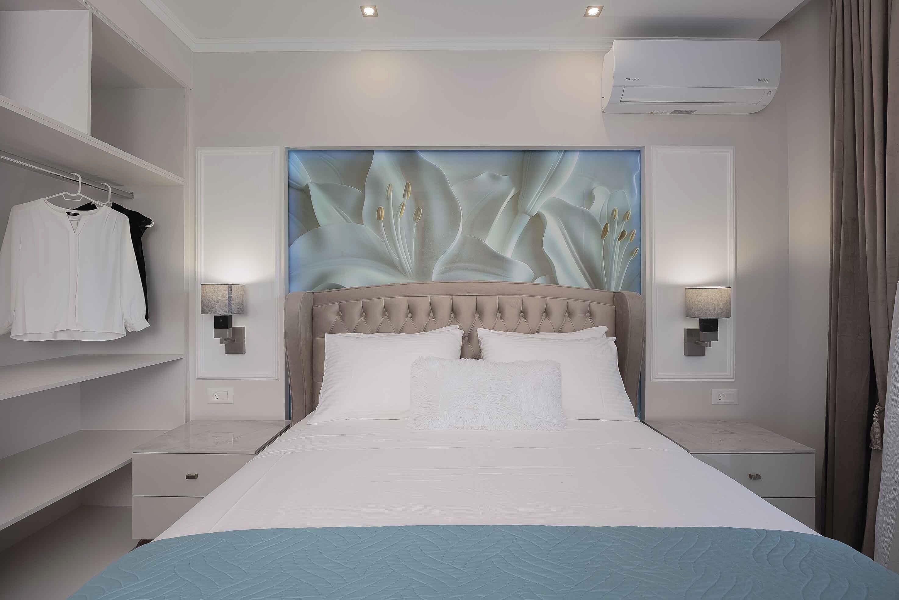 A modern hotel room with a tufted headboard, a large wall mural, a bed with white bedding, and a wardrobe with hanging clothes.
