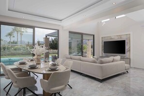 A modern living room with a dining area, large windows, a flat-screen TV, and a pool view. - Atriora Luxury Villa | Private Pool & 150m to sea! (Kissamos)