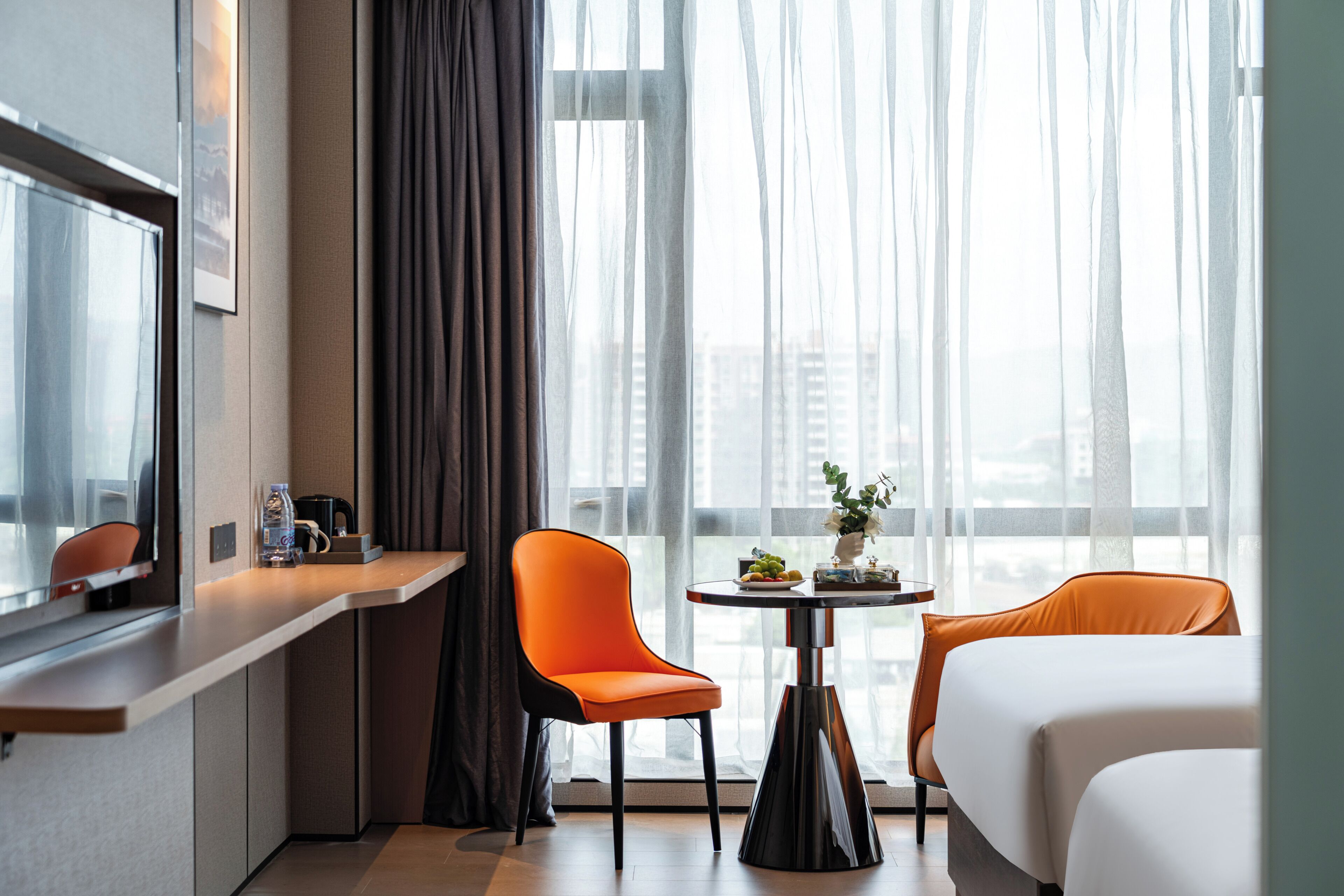 A hotel room with a bed, a desk with a chair, a small table with a vase, and a large window with sheer curtains.