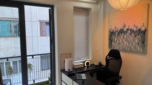 Ein modernes Schlafzimmer mit einem Bett, einem Schreibtisch mit Computer, einem Sessel, einem Fenster mit Jalousien und einer Pendelleuchte.