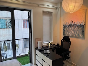A modern bedroom with a bed, a desk with a computer, a chair, a window with blinds, and a hanging light fixture. - Cozy Studio Apartment in Prime District One, Ho Chi Minh City (Thành phố Hồ Chí Minh)