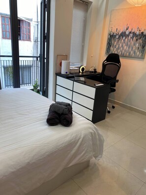 A bedroom with a bed, a desk with a chair, a window with blinds, and a painting on the wall. - Cozy Studio Apartment in Prime District One, Ho Chi Minh City (Thành phố Hồ Chí Minh)