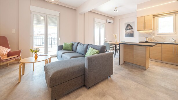 Ein modernes Wohnzimmer mit einem grauen Ecksofa, einem kleinen runden Tisch und einem roten Sessel. Es gibt eine Küchenzeile mit Holzschränken und einem Fenster über der Spüle.