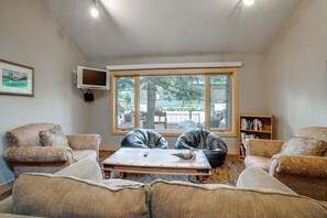 A living room with a large window, a flat-screen TV mounted on the wall, and a bookshelf. - Keystone Lakefront Retreat! Pool & Hot Tub Access (Keystone)