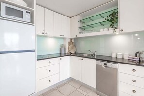 A modern kitchen with white cabinets, a microwave, a dishwasher, and a glass shelf with kitchenware. - Noosa Luxury on Hastings St w/ Pool, Spa & Gym (Noosa)