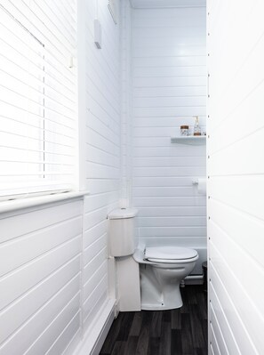 Ein kleines Badezimmer mit weißen Wänden, einer Toilette, einem Fenster und einem Ablagefach für Pflegeprodukte.
