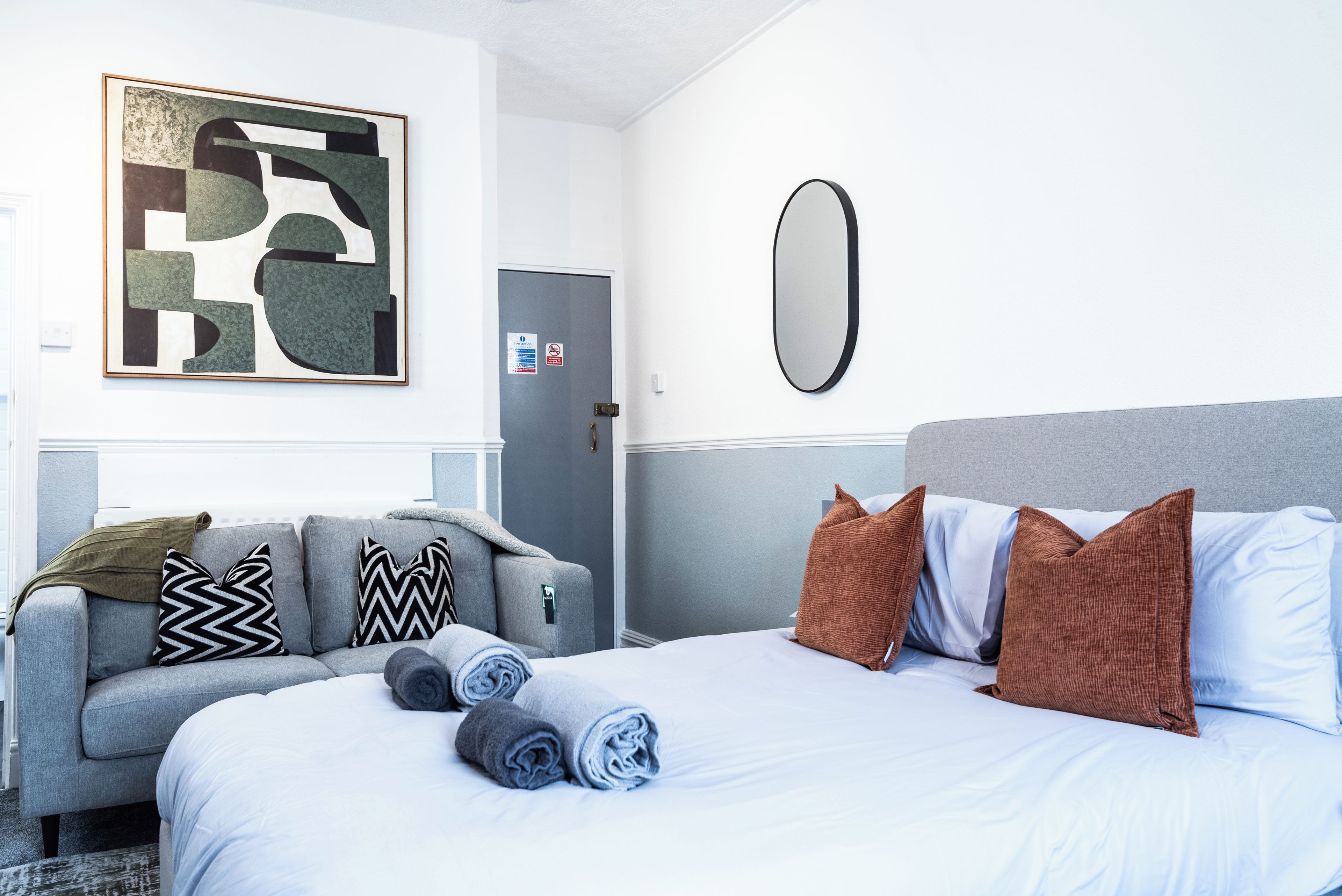 A hotel room with a bed, a grey sofa, a wall-mounted mirror, and a painting.