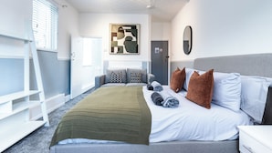 Een hotelkamer met een bed, kussens, dekens, een nachtkastje, een lamp, een raam met jaloezieën, een deur en een kunstwerk aan de muur.