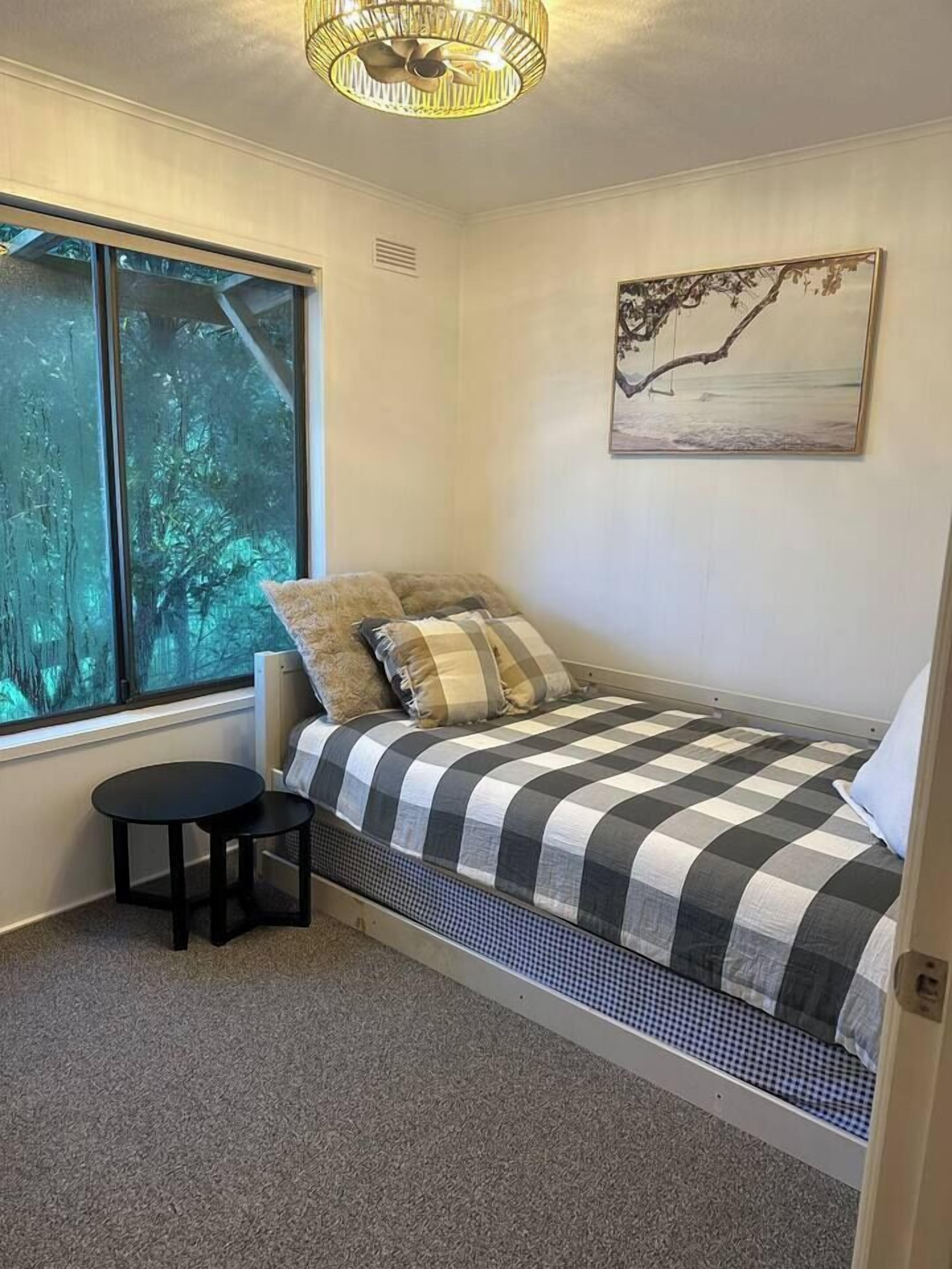 Une petite chambre à coucher avec un lit, une table ronde, une fenêtre donnant sur des arbres et un tableau accroché au mur.