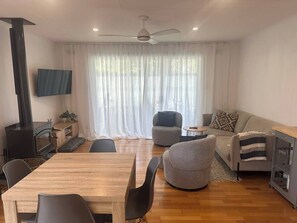 A modern living room with a wooden dining table, two armchairs, a sofa, a TV mounted on the wall, and a fireplace.