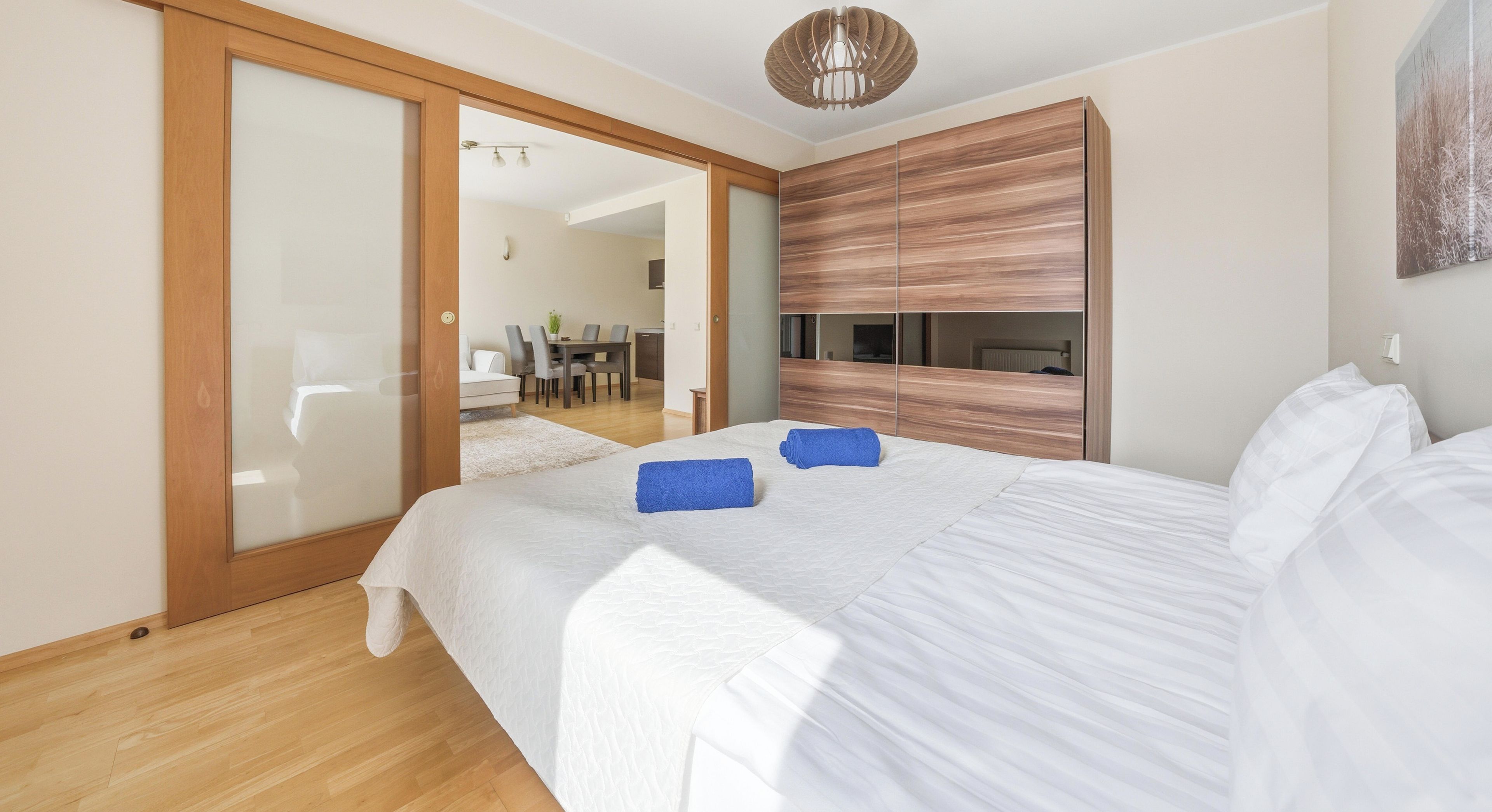 Een slaapkamer met een groot bed, een houten garderobe, een schuifdeur naar een badkamer, en een eethoek die door de deur zichtbaar is.