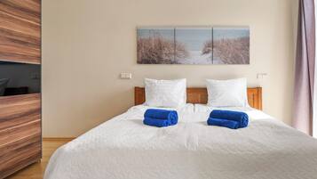 Hotellihuone, jossa on sänky, valkoiset vuodevaatteet, kaksi tyynyä, sininen pyyhe, puinen pääty, taulu seinällä ja puulattia.