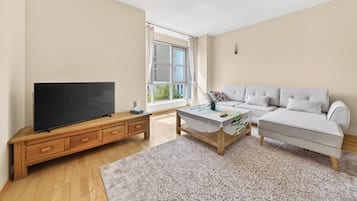 Een moderne woonkamer met een flatscreen televisie op een houten tv-meubel, een bank, een salontafel met een geruit tafelkleed en een groot raam met gordijnen.