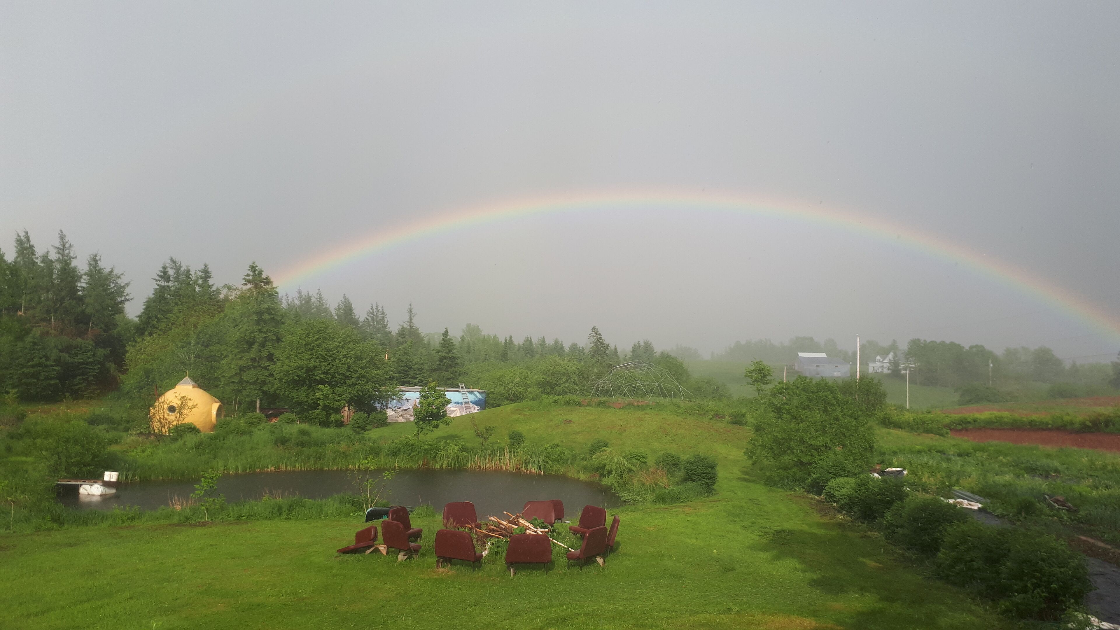 A rural landscape with a rainbow, a pond, and a cluster of red chairs.