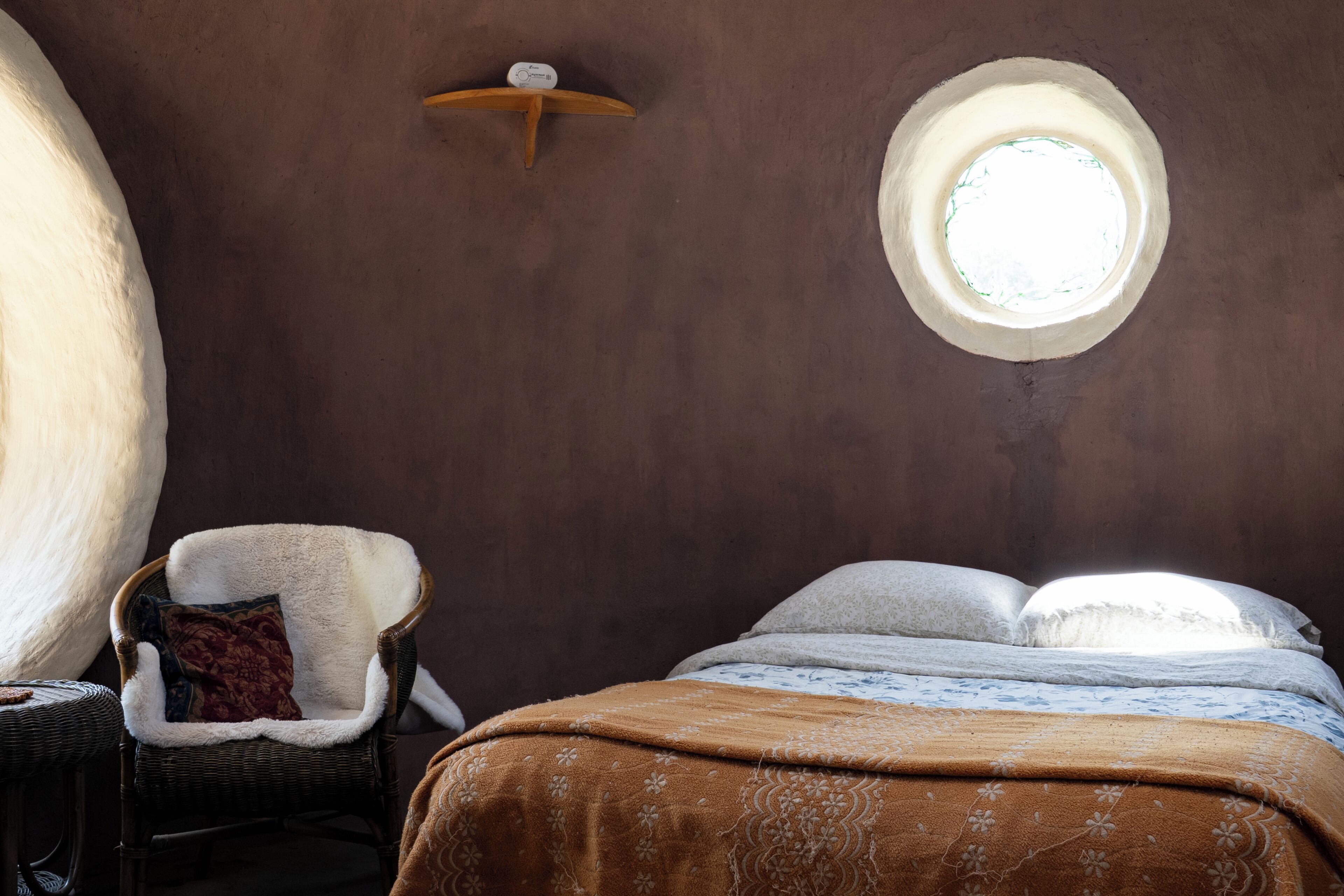 Une pièce chaleureuse avec une fenêtre ronde, un fauteuil en osier doté d’un coussin et un lit recouvert d’une couverture à motifs.