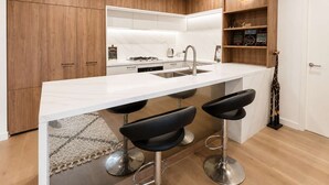 Una cucina moderna con un piano di lavoro in marmo bianco, elettrodomestici in acciaio inossidabile e mobili in legno. Sotto l'isola si trovano tre sgabelli da bar neri.