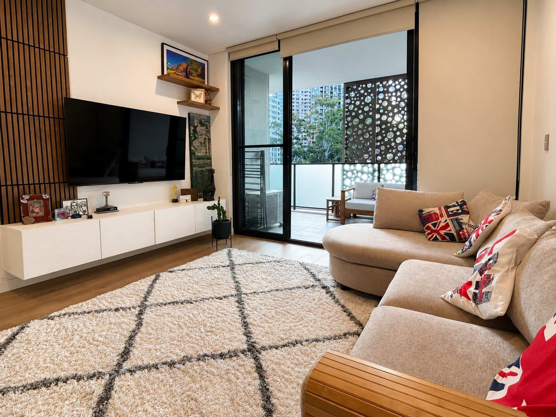 A modern living room with a flat-screen TV, a sofa with patterned cushions, a wooden sideboard, and a large window with a view of buildings and greenery.