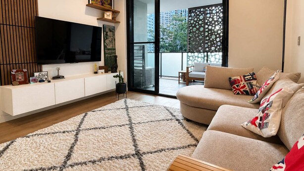 Un soggiorno moderno con una televisione a schermo piatto, un divano con cuscini decorati, un mobiletto in legno e un'ampia finestra vista edifici e vegetazione.