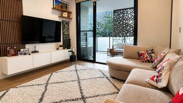 A modern living room with a flat-screen TV, a sofa with patterned cushions, a wooden sideboard, and a large window with a view of buildings and greenery.