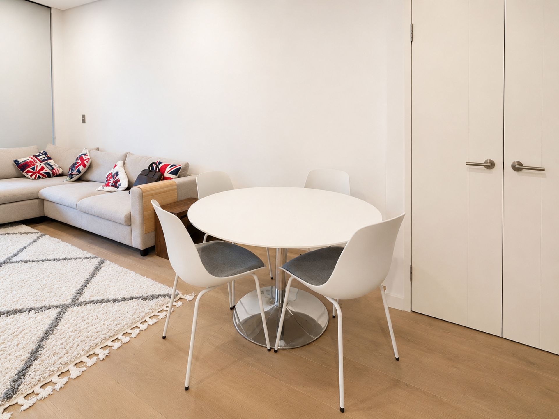 A modern living room with a round white table, two white chairs, a beige sofa with patterned cushions, and a white door.