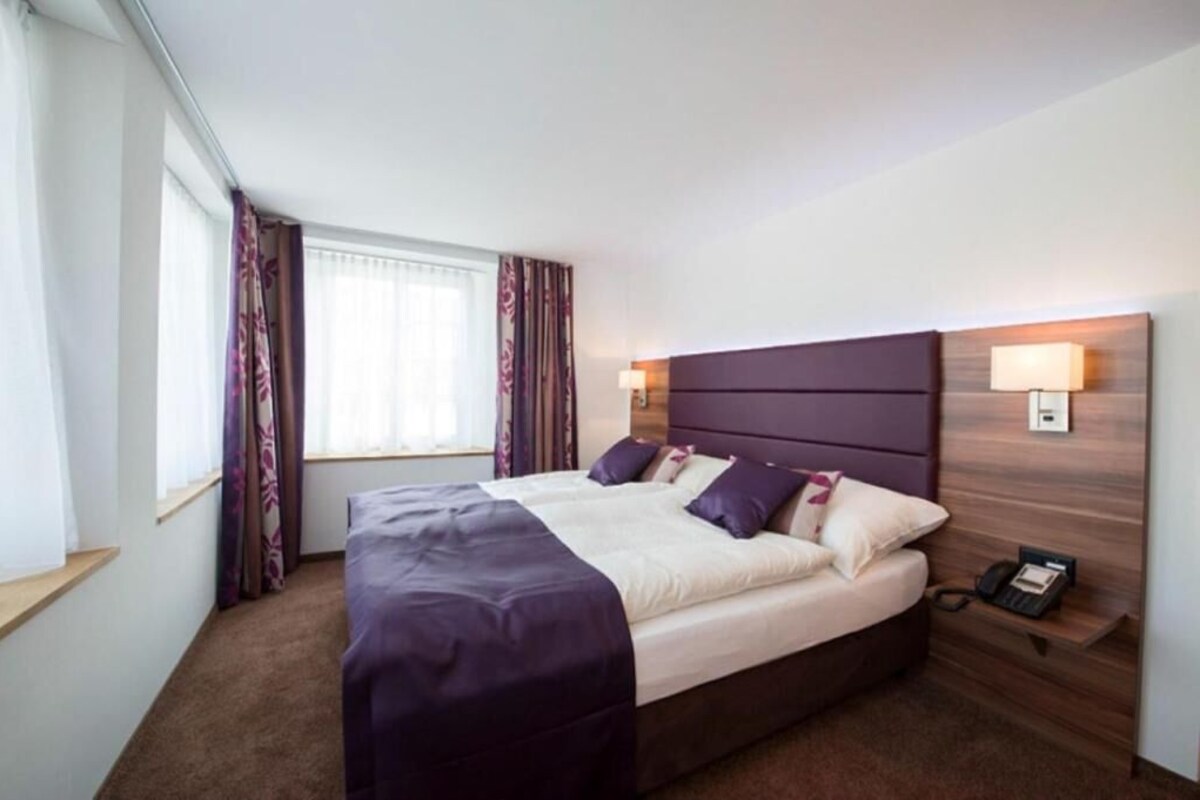 A hotel room with a large bed, bedside lamps, a nightstand, and a window with curtains.