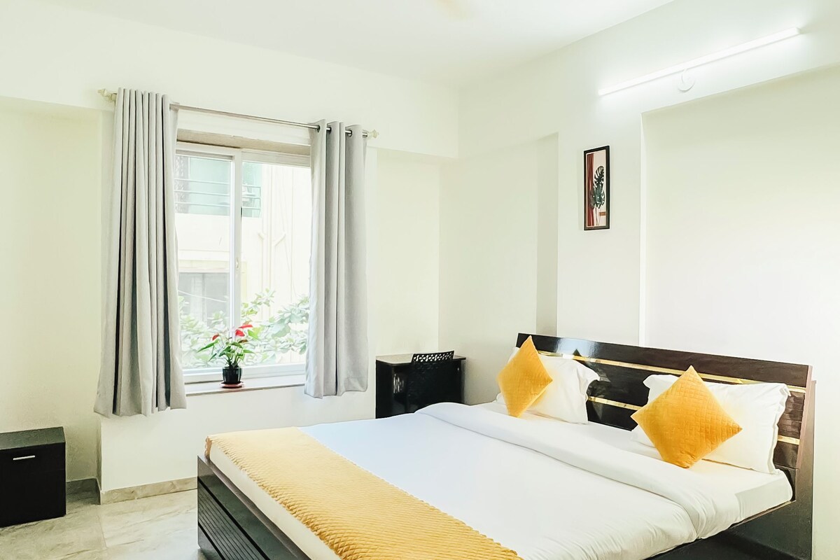 A bedroom with a bed, a ceiling fan, a nightstand, a bedside table, a window with curtains, and a small potted plant.