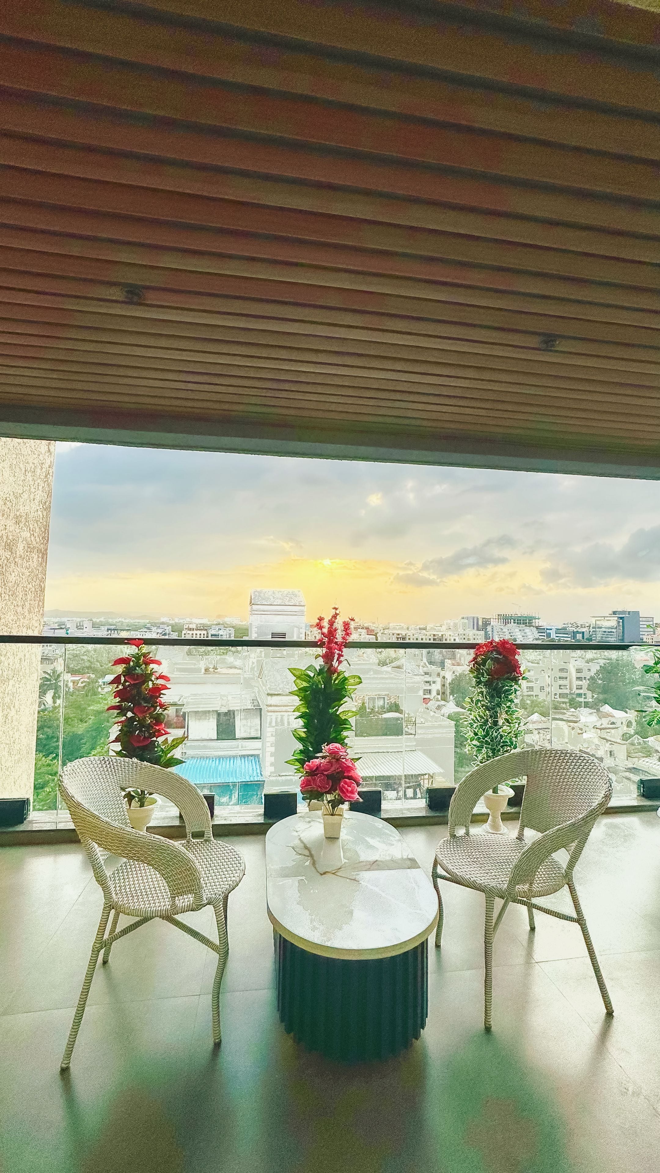 Un balcón con sillas de mimbre y una mesa, con vistas a un paisaje urbano con edificios y una puesta de sol.