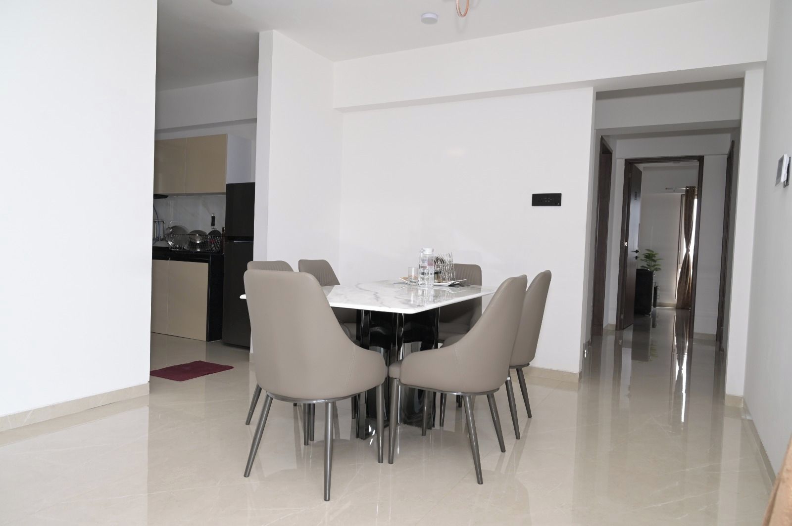 A modern dining area with a white table and four chairs, a kitchen in the background, and a hallway leading to another room.